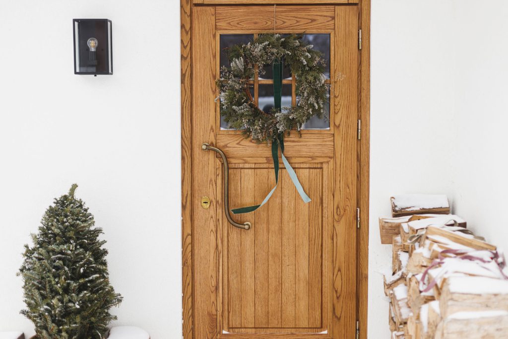 Minimalist Christmas door decorations of a simple wreath on a wooden door with a small Christmas tree next to the door