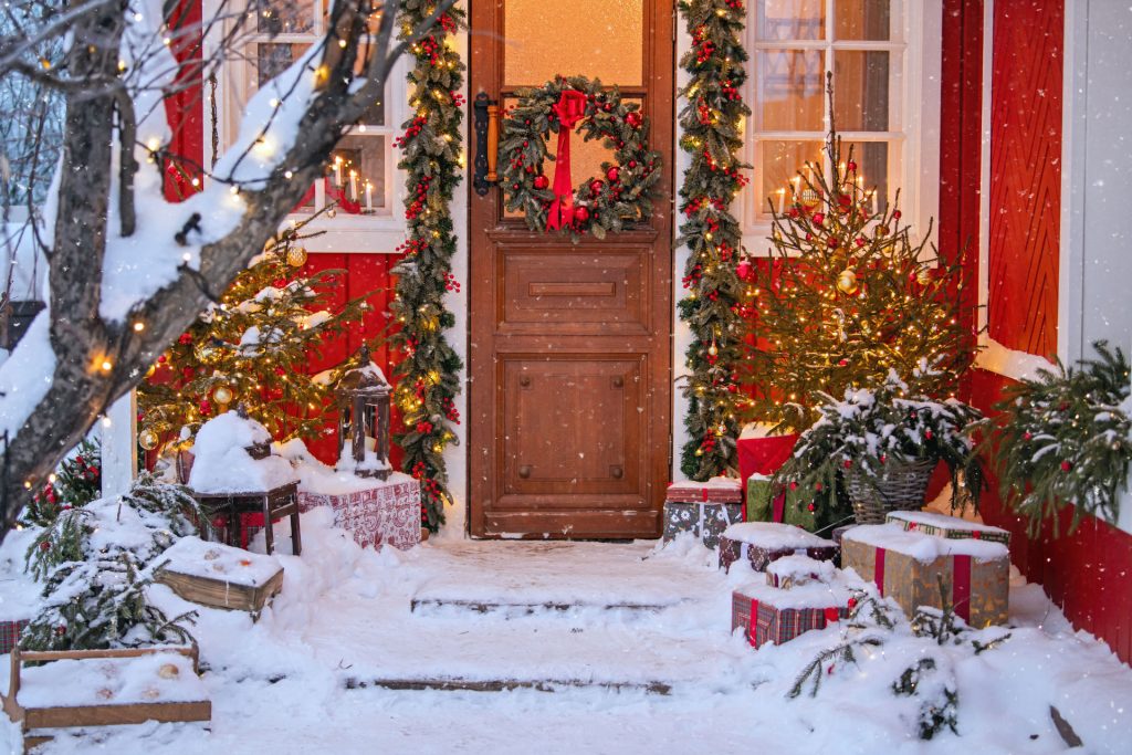 Front porch with Christmas lights, candles, garlands, wreaths, Christmas trees, and presents in falling snow