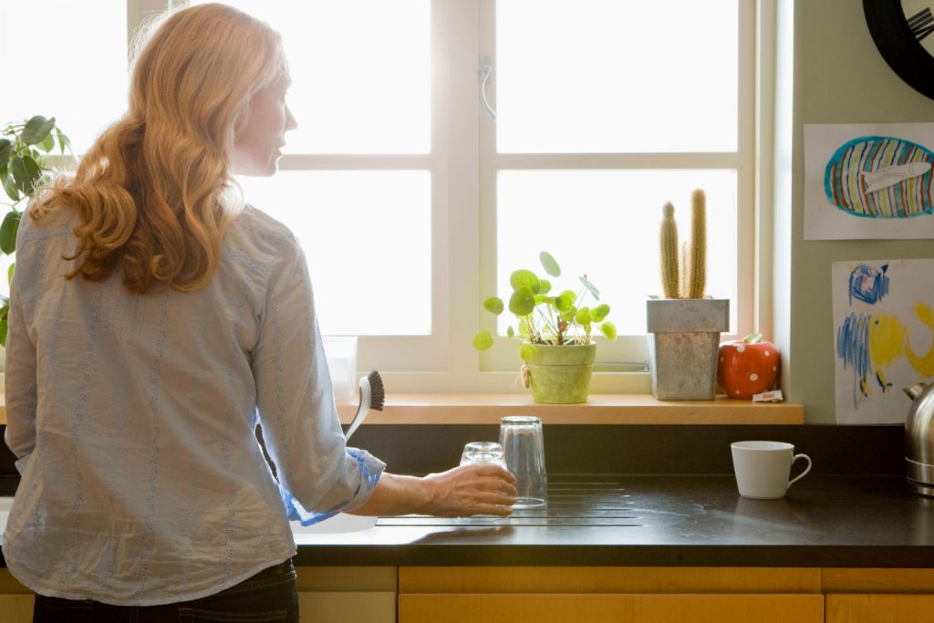 A woman sets a drinking glass on the kitchen counter while peering out the kitchen sink window