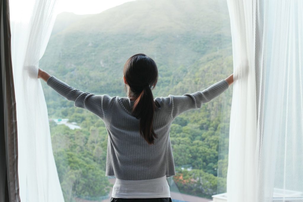 A woman spreads open the sheer white window curtains