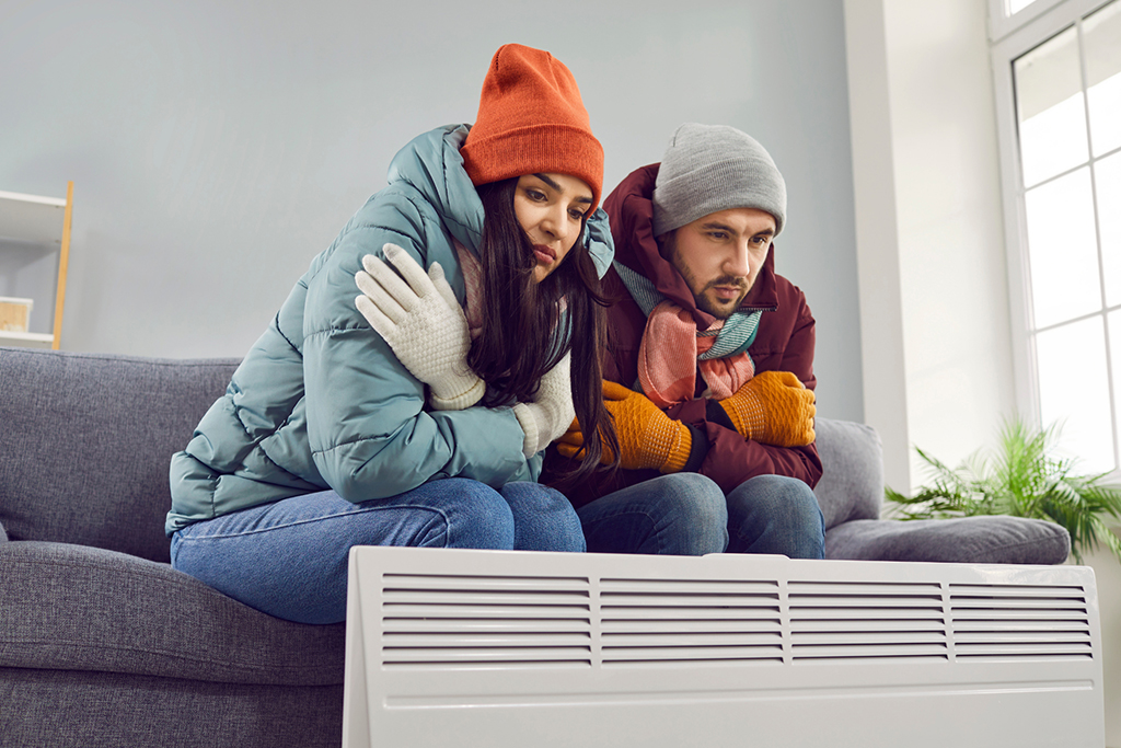 A young couple in winter apparel sits shivering in their home